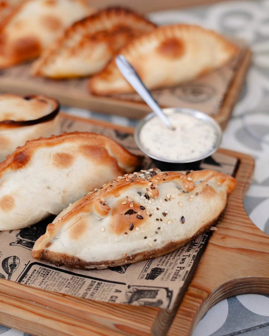 Empanadas douradas, uma com sementes de sésamo, sobre tábuas de madeira com papel de jornal, ao lado de um recipiente com molho branco e uma colher