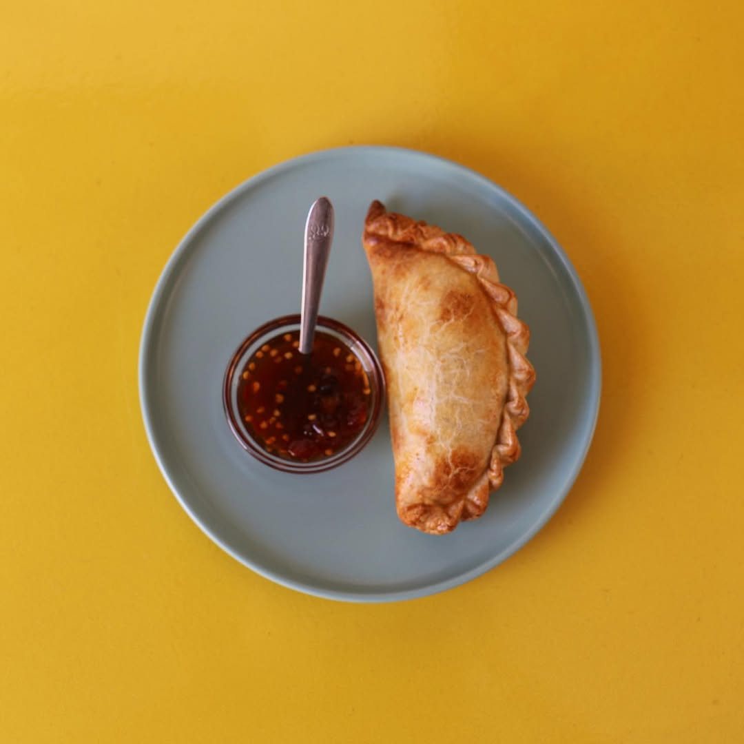 Uma empanada dourada num prato cinzento, ao lado de uma taça pequena com molho vermelho e uma colher, sobre fundo amarelo